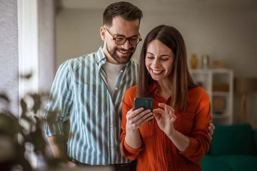 Cheerful young couple smiling together at home, looking at phone