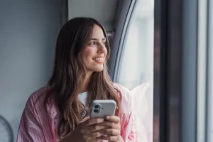 A smiling young woman holds her smartphone and gazes out a window